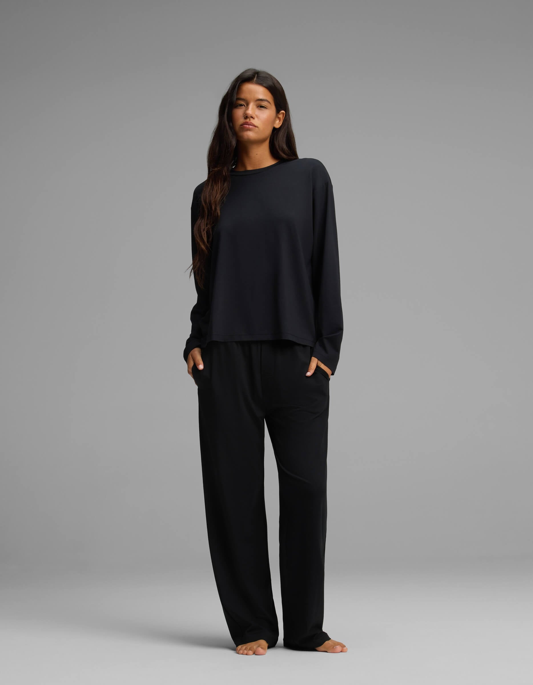 A woman with long dark hair stands barefoot against a gray background, wearing Cozy Earth’s Women’s First Layer Long Sleeve Sleep Tee and wide-leg black pants, with her hands in her pockets.
