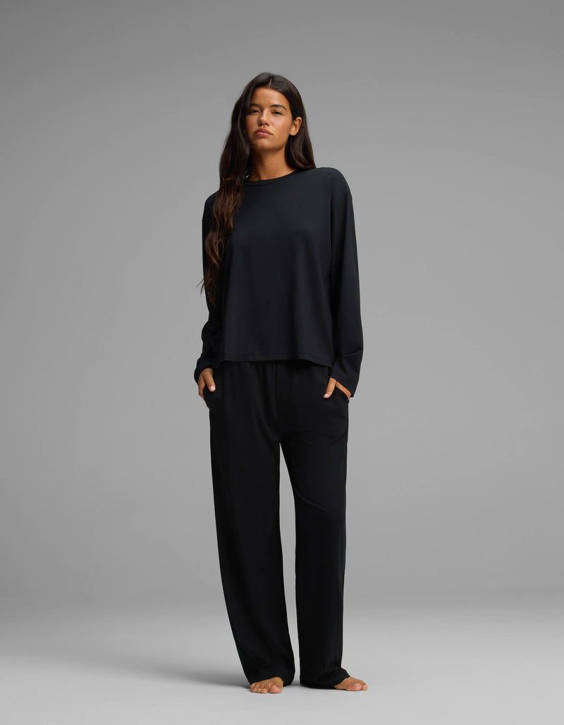A woman with long dark hair stands barefoot against a gray background, wearing Cozy Earth’s Women’s First Layer Long Sleeve Sleep Tee and wide-leg black pants, with her hands in her pockets.