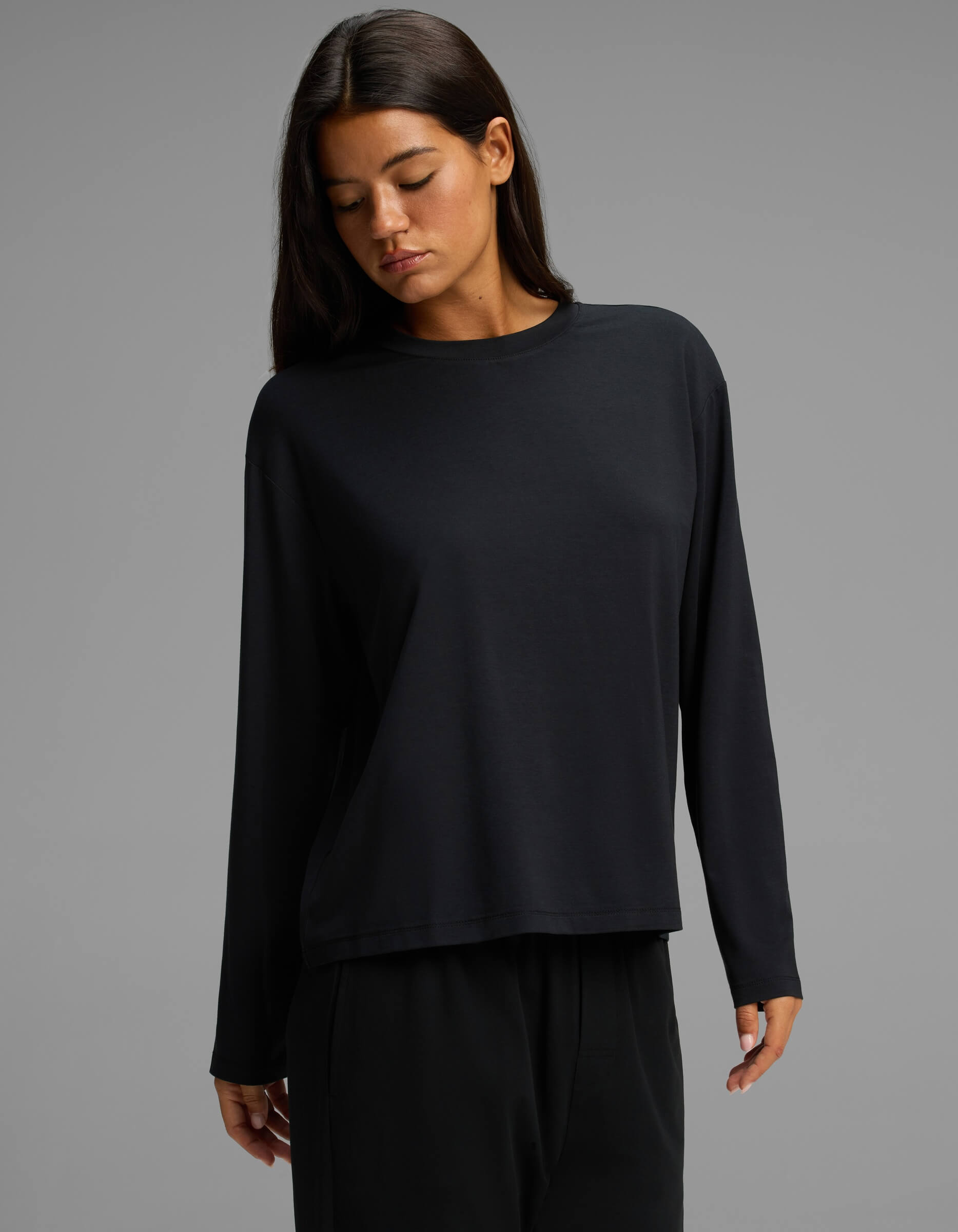 A model with long, dark hair gazes downward while wearing the Cozy Earth Women’s First Layer Long Sleeve Sleep Tee & Pajama Pant Set in black, set against a neutral gray background.