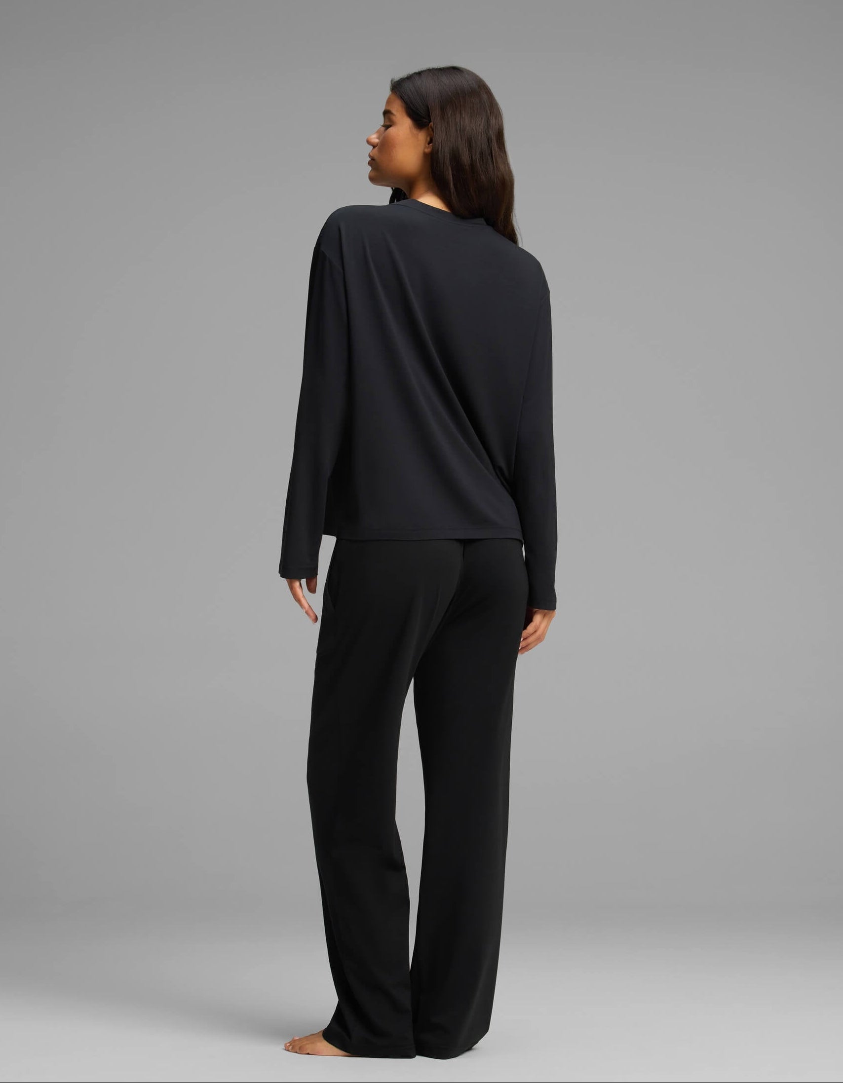 A woman with long brown hair, seen from behind, stands barefoot against a plain gray background wearing the Cozy Earth Women’s First Layer Long Sleeve Sleep Tee & Pajama Pant Set in black.