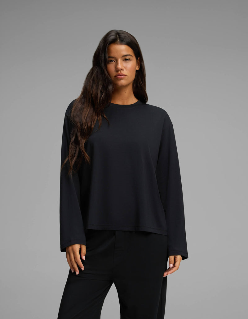 A woman with long dark hair wears the Cozy Earth Women’s First Layer Long Sleeve Sleep Tee in black, paired with black pants, standing against a plain gray background.