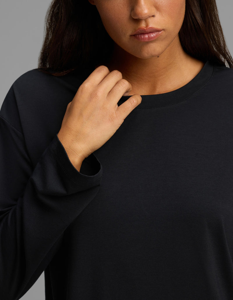 A woman wearing the Cozy Earth Women’s First Layer Long Sleeve Sleep Tee in black touches her collar with her right hand. Only the lower half of her face, neck, and upper torso are visible against a neutral gray background.