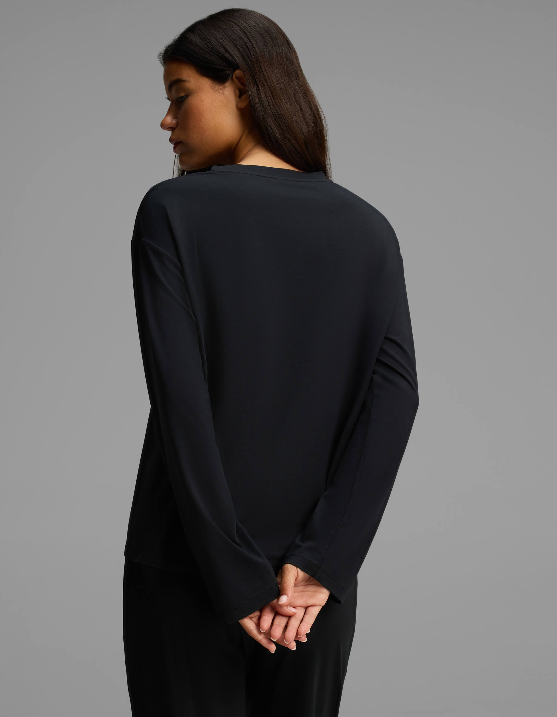 A woman with long dark hair stands facing away, her head turned to the side. She is wearing Cozy Earth’s Women’s First Layer Long Sleeve Sleep Tee in black, paired with black pants, her hands clasped behind her back against a plain gray background.