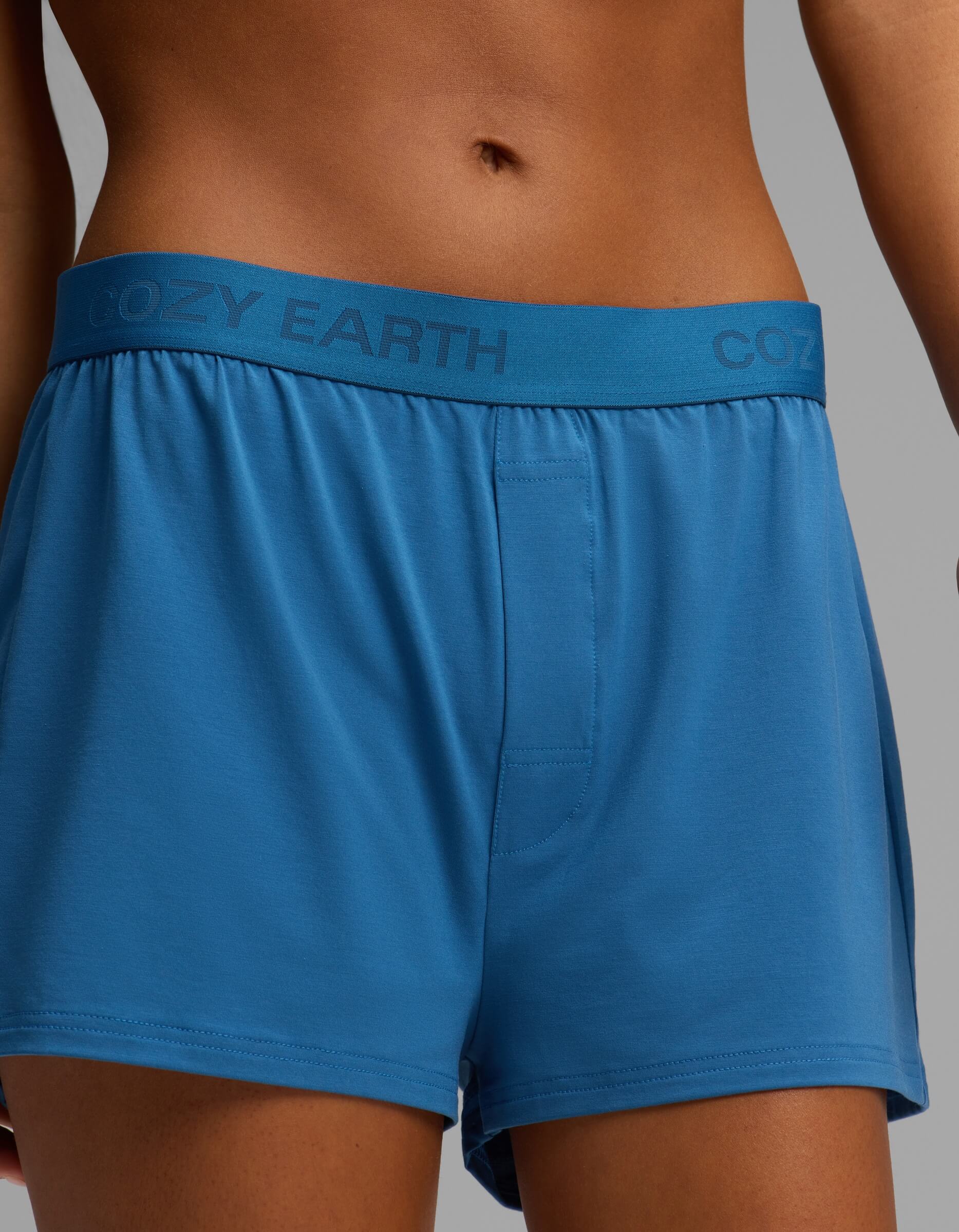A person models the Cozy Earth Women’s First Layer Boyfriend Sleep Tee & Boxer Short Set, featuring blue boxer shorts with the brand name “Cozy Earth” printed on the waistband, against a neutral background. Only the torso and upper legs are visible.