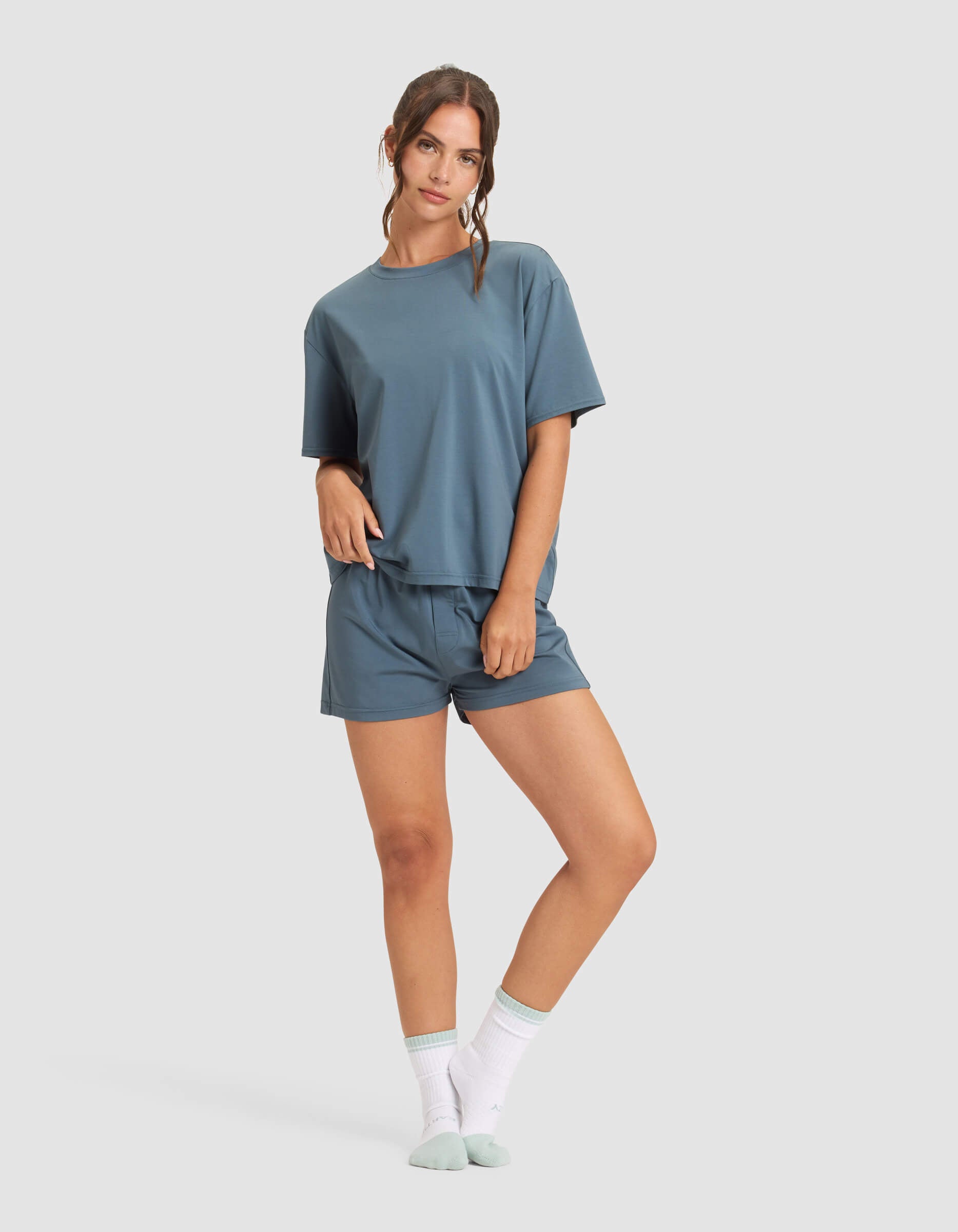 A woman poses casually in the Cozy Earth Women's First Layer Pajama Short Sleeve Tee & Boxer Short Set, featuring a loose fit in blue, styled with white crew socks and light blue slippers against a plain background.