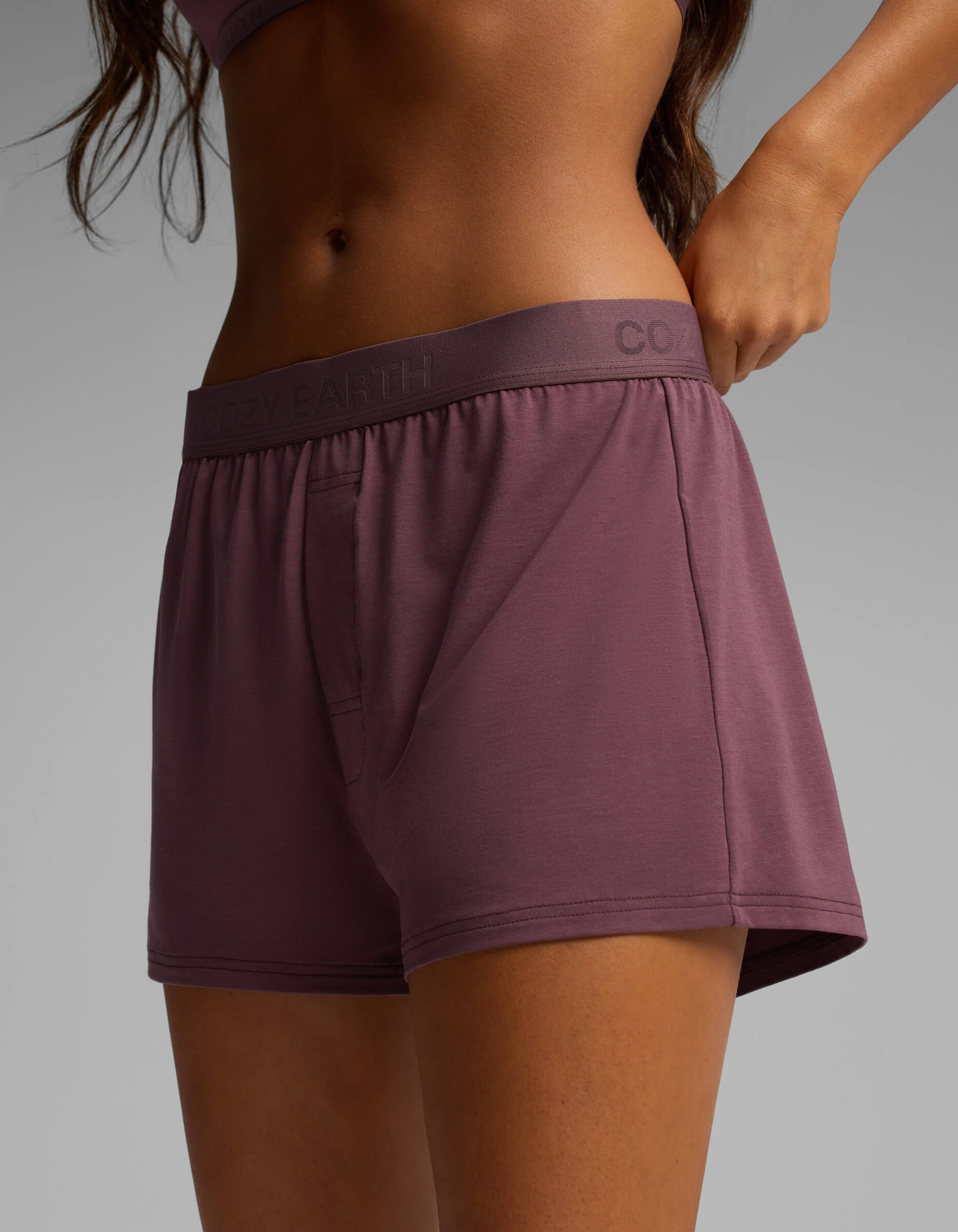 A person wearing the mauve-colored Women’s First Layer Boyfriend Sleep Tee & Boxer Short Set from Cozy Earth, shown from mid-torso to upper thigh, stands against a plain gray background.