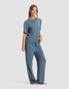 A woman stands barefoot against a plain background, wearing the Cozy Earth Women's First Layer Pajama Short Sleeve Tee & Pant Set. Her wavy hair is tied back, and she has a relaxed expression.|Color:Dawn