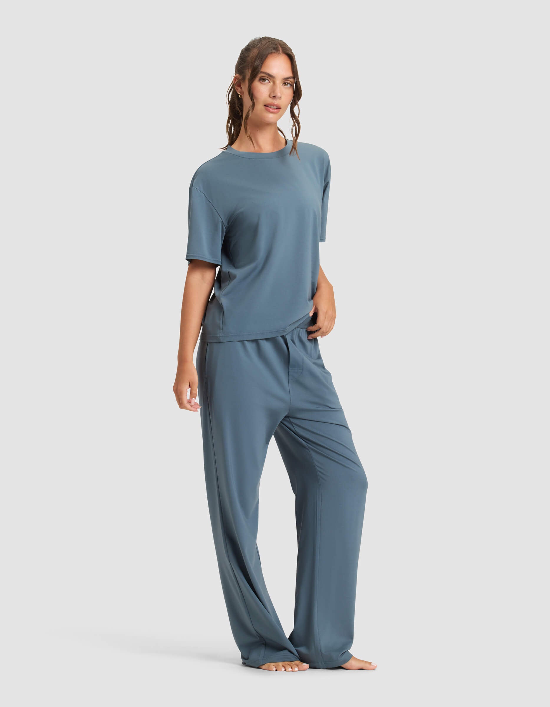 A woman stands barefoot against a plain background, wearing the Cozy Earth Women's First Layer Pajama Short Sleeve Tee & Pant Set. Her wavy hair is tied back, and she has a relaxed expression.|Color:Dawn