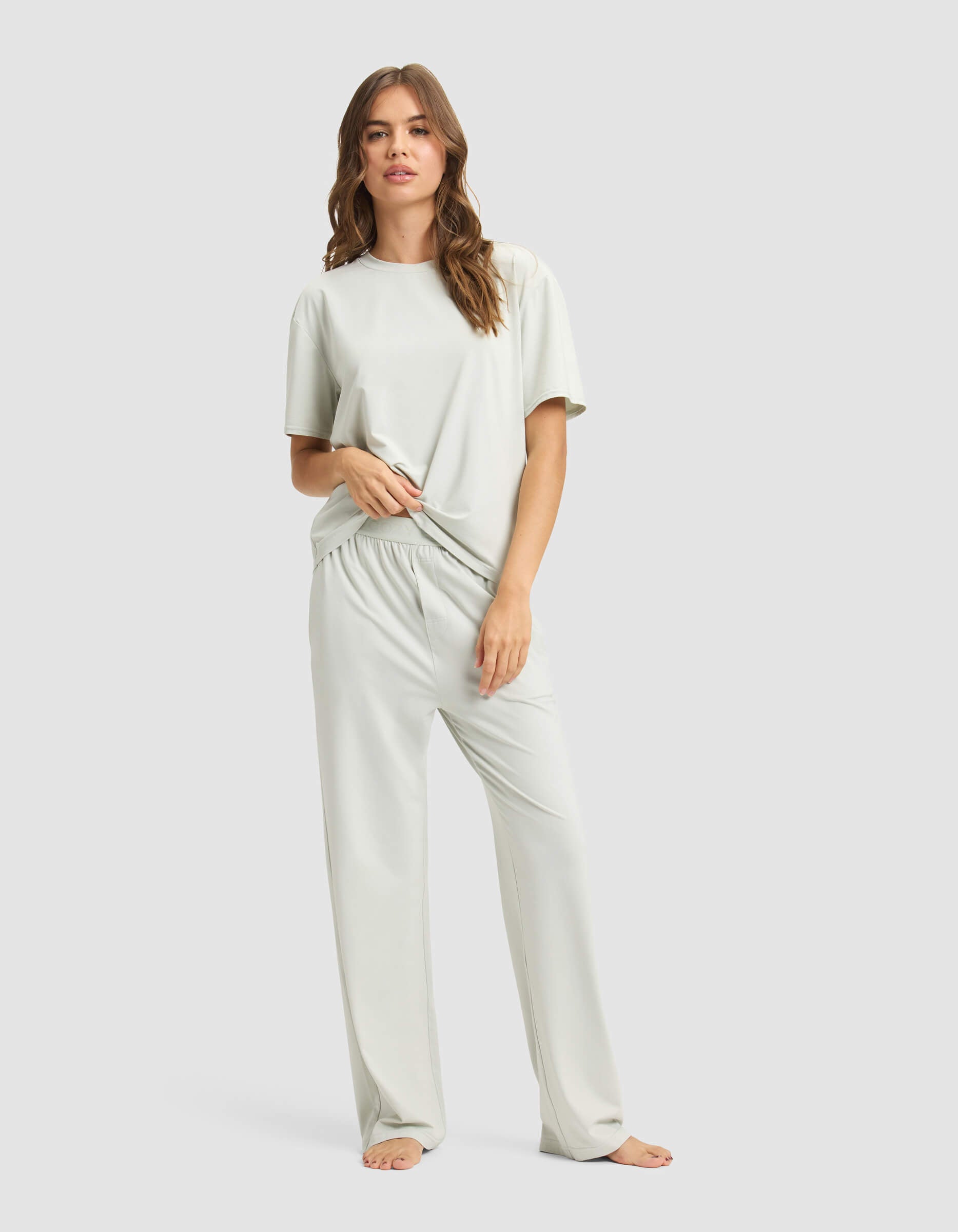 A woman stands barefoot against a plain background, wearing the Cozy Earth Women's First Layer Pajama Short Sleeve Tee & Pant Set in a loose fit, holding part of her shirt with one hand.|Color:Laurel