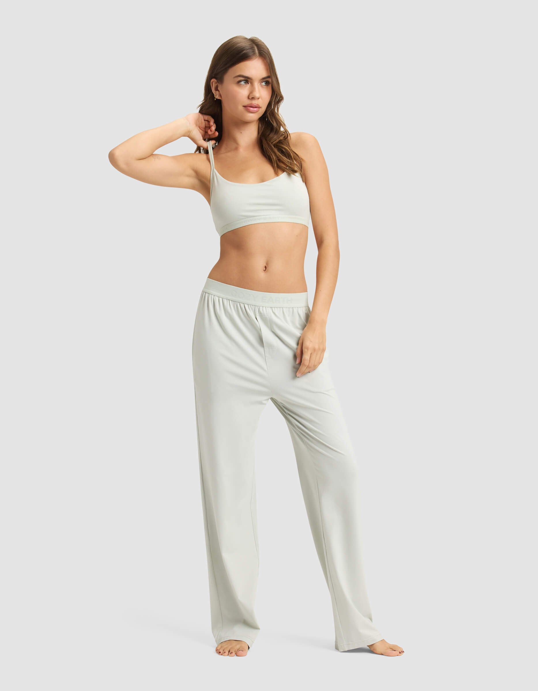 A woman with long wavy hair stands barefoot against a plain background, wearing the Cozy Earth Women's First Layer Pajama Soft Bra & Pant Set in light gray, with one hand resting on her head.|Color:Laurel