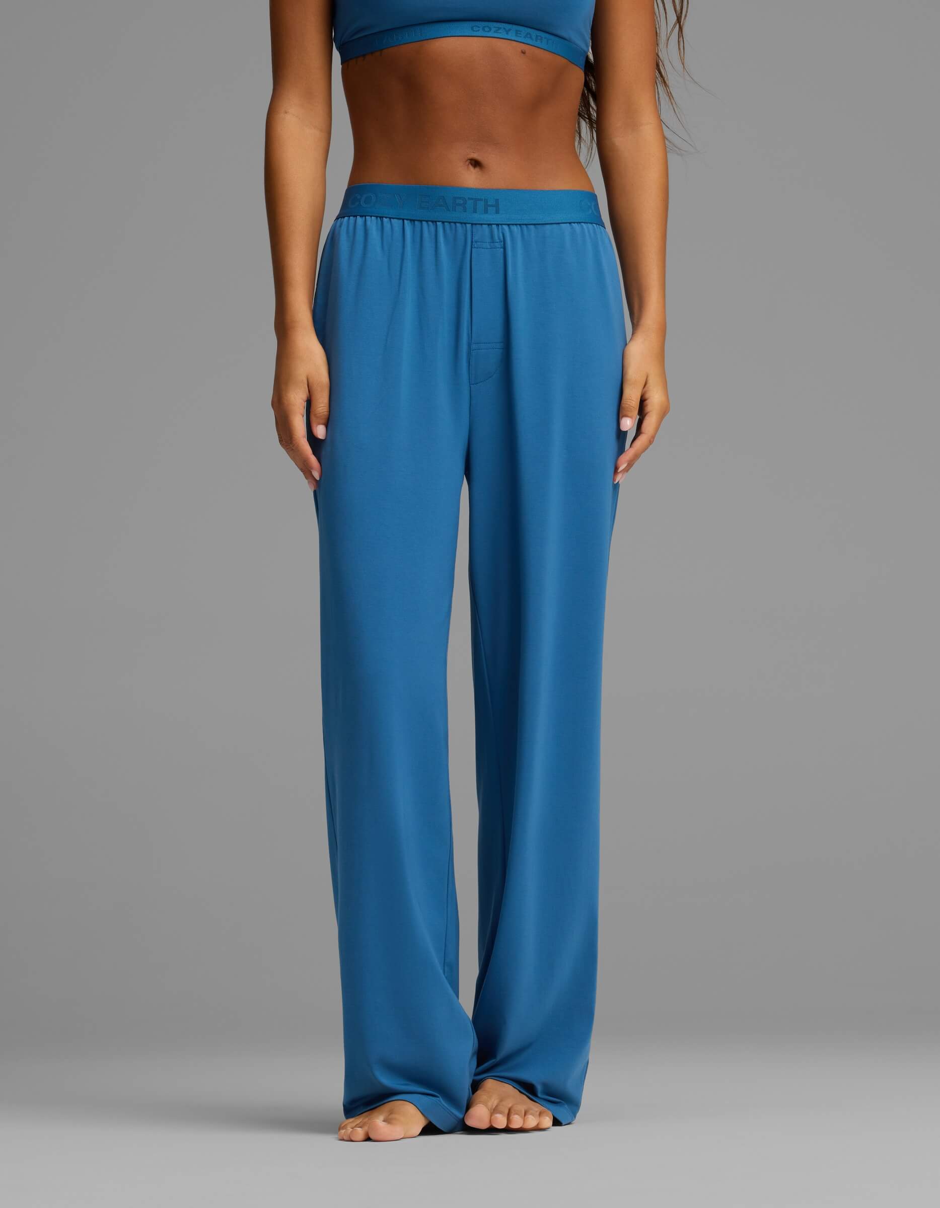 A person stands barefoot, wearing Cozy Earth’s Women’s First Layer Pajama Pant in blue with a matching top. The image is cropped from the shoulders down, with hands resting at the sides against a plain gray background.|Color:Anchor