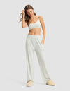 A woman in Cozy Earth's Women's First Layer Pajama Soft Bra & Pant Set stands against a plain background, wearing the light bralette and loose pajama pants with beige slippers, one hand in her pocket and the other touching her hair.|Color:Laurel