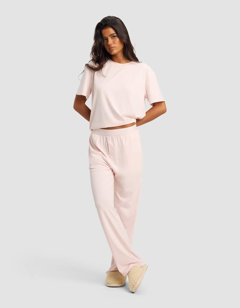 A woman models the Cozy Earth Women's First Layer Pajama Short Sleeve Tee & Pant Set in light pink with beige slippers, standing against a plain light gray background.