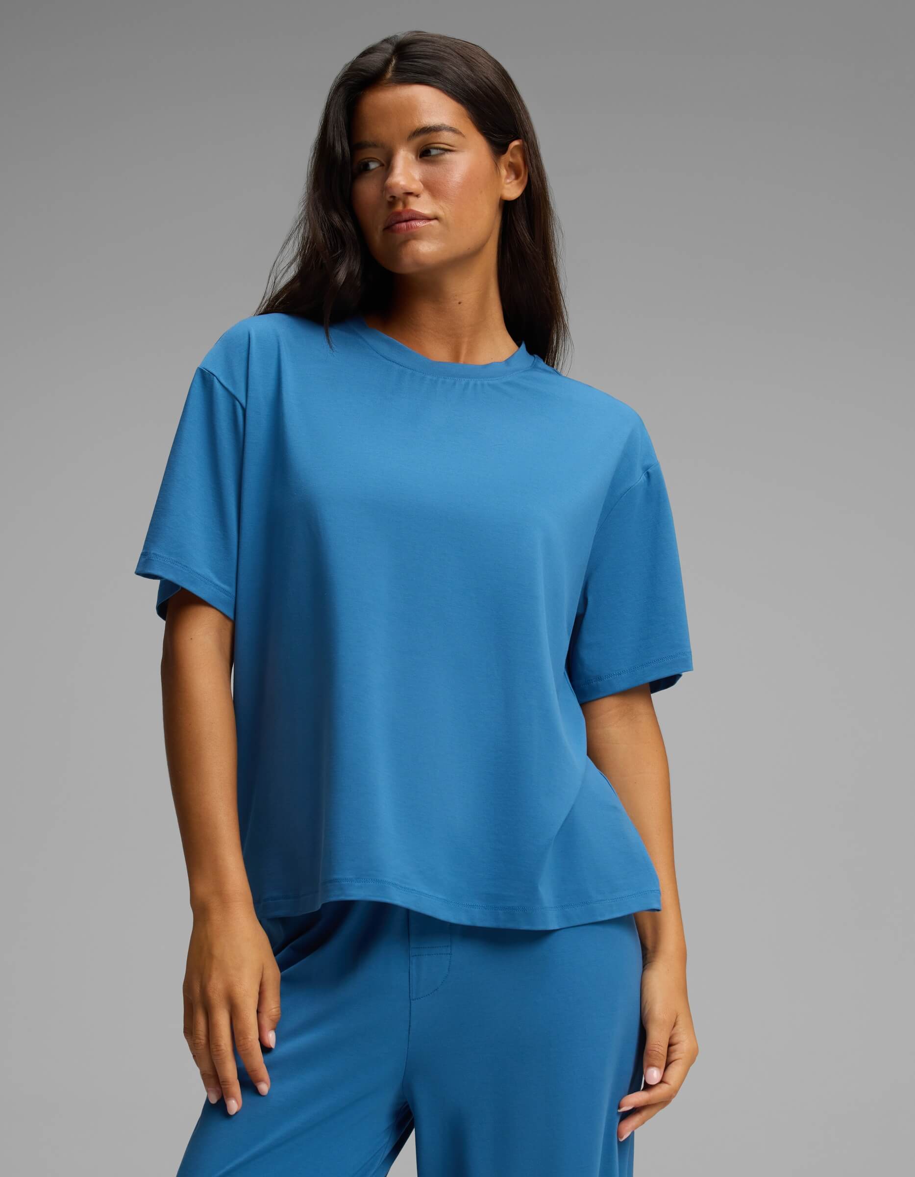 A woman with long dark hair wears the Cozy Earth Women’s First Layer Pajama Short Sleeve Tee in blue, paired with matching blue pants. She stands against a plain gray background, looking slightly to the side with a neutral expression.|Color:Anchor