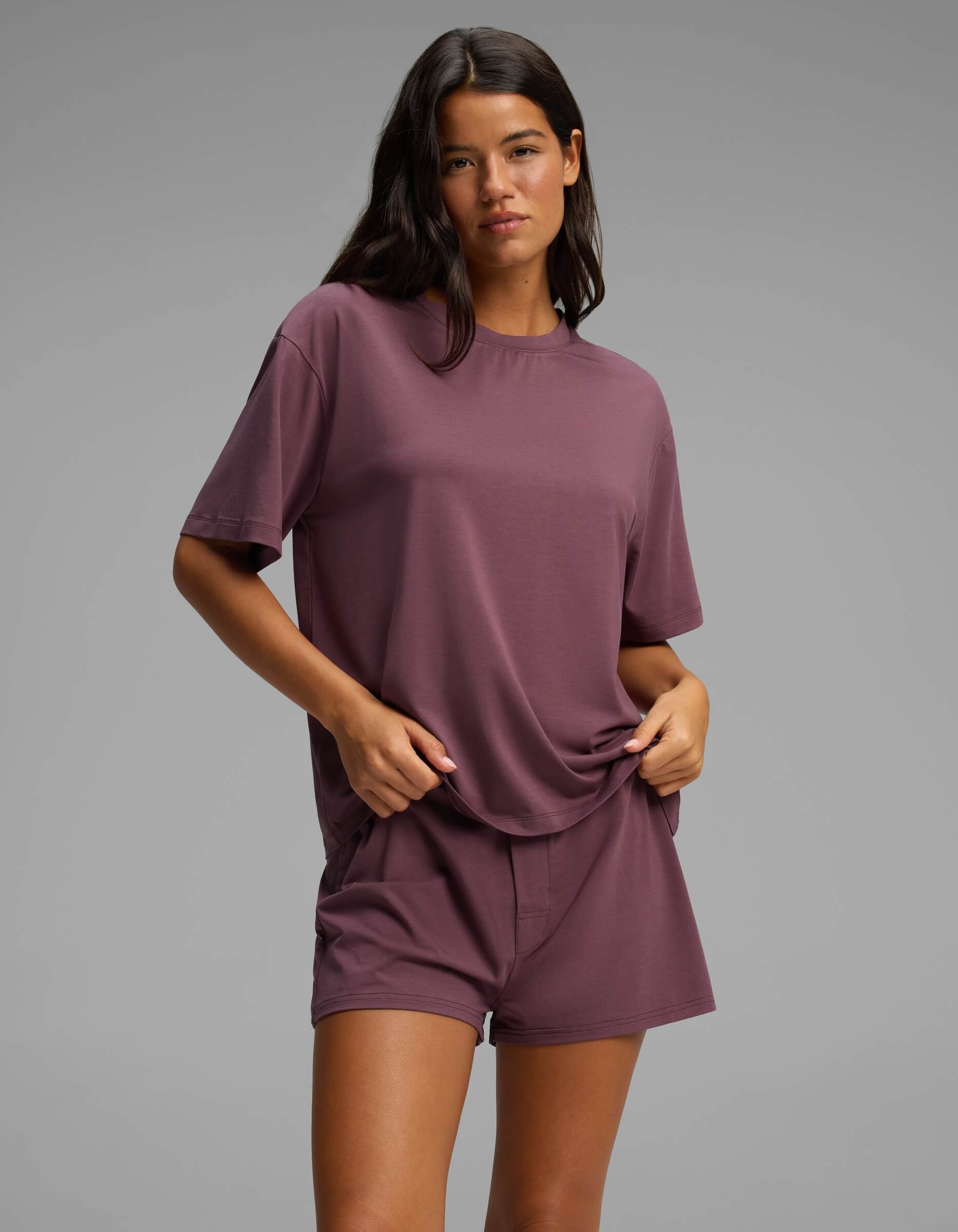 A woman stands against a plain gray background, wearing the Cozy Earth Women’s First Layer Pajama Short Sleeve Tee in mauve with matching shorts, one hand holding her shirt and the other in her pocket.|Color:Fig