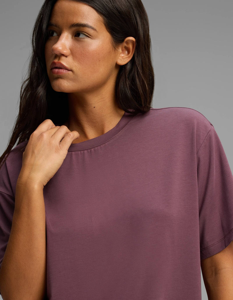 A woman with long dark hair wearing the Cozy Earth Women’s First Layer Pajama Short Sleeve Tee in mauve looks to the side against a plain gray background.
