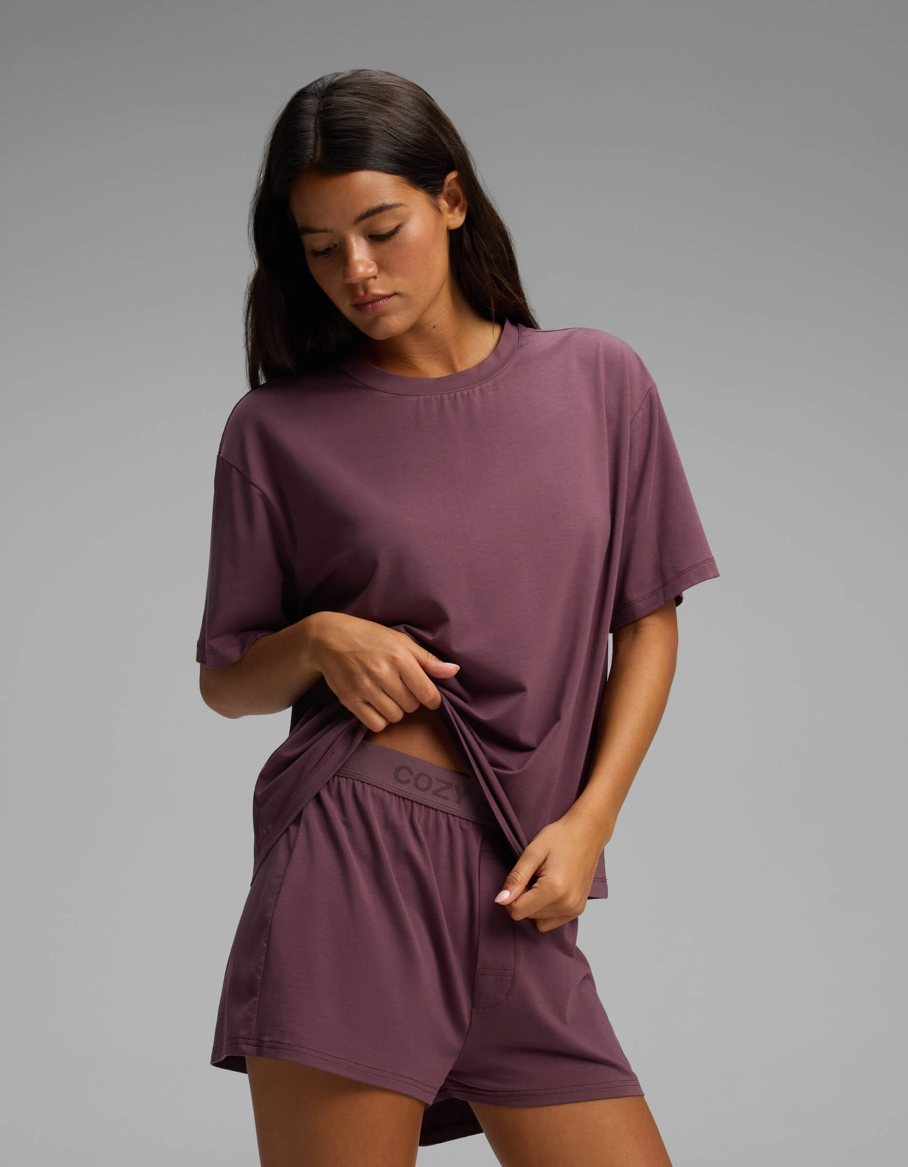 A woman stands against a plain background, slightly lifting the hem of her shirt to reveal the waistband of her shorts, while wearing the Cozy Earth Women’s First Layer Pajama Short Sleeve Tee & Boxer Short Set in a matching mauve color.