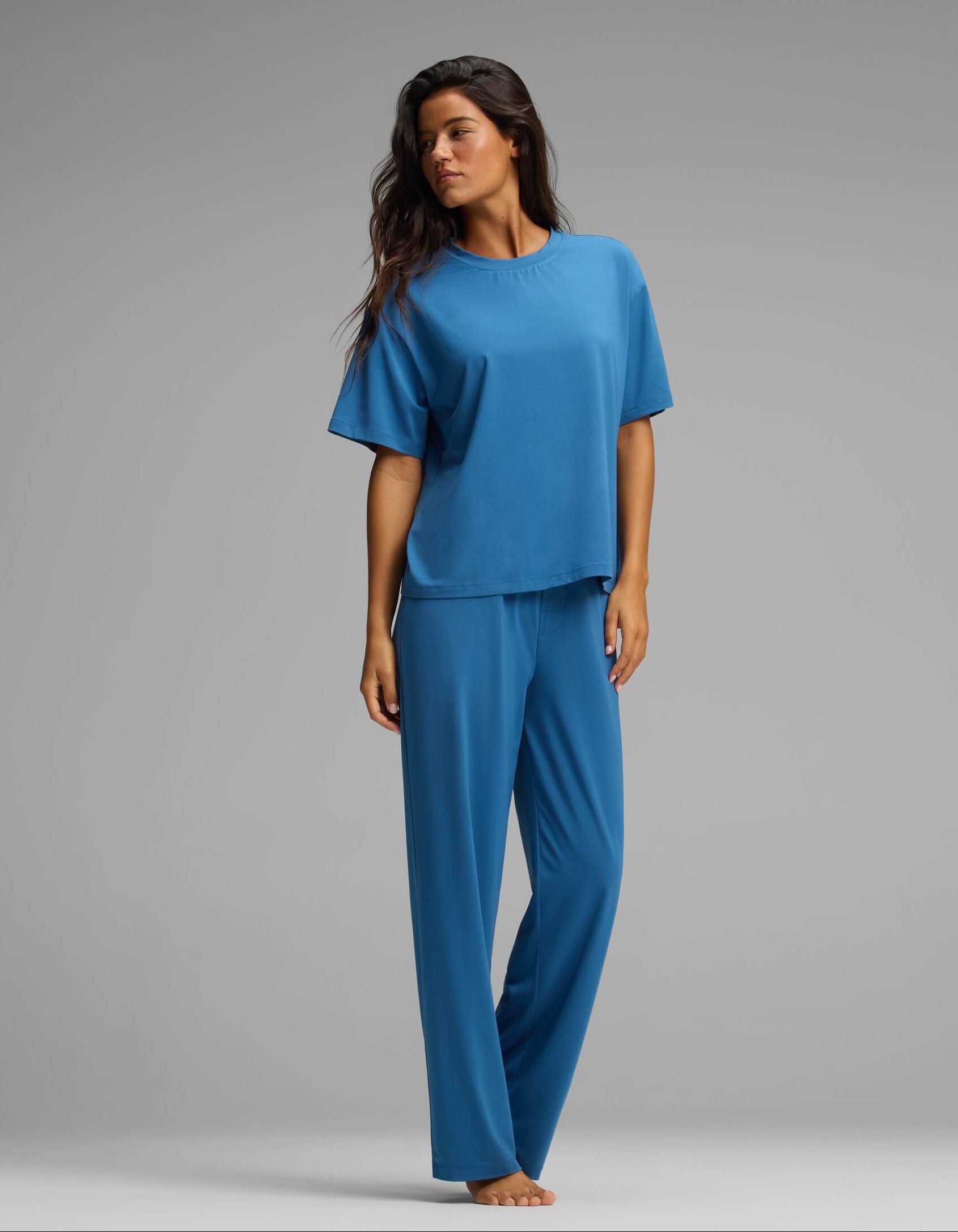 A woman stands barefoot against a gray background, wearing the Cozy Earth Women’s First Layer Pajama Short Sleeve Tee & Pant Set in a relaxed, loose-fitting style, looking to the side with an easygoing posture.|Color:Anchor