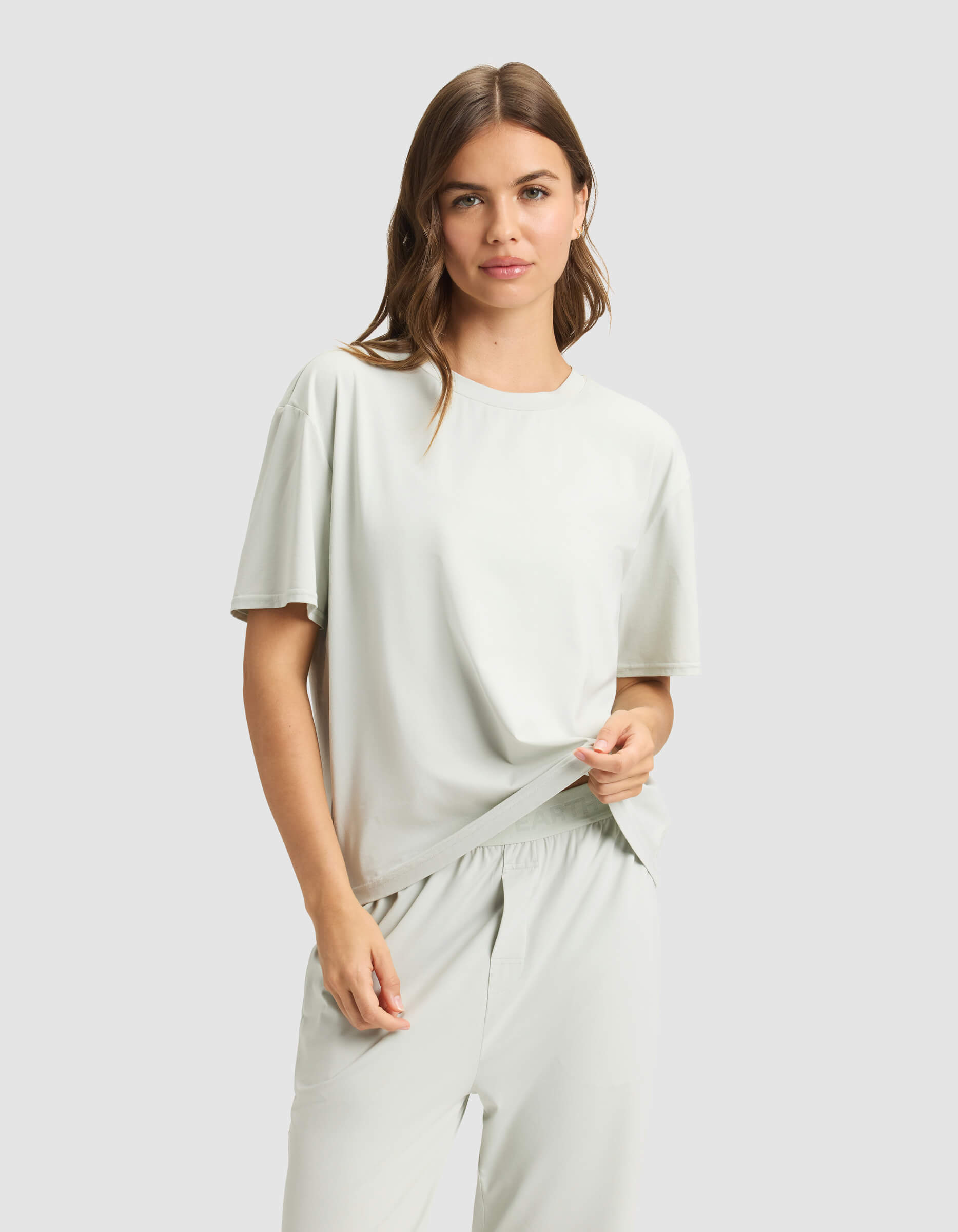 A woman with long brown hair wears the Cozy Earth Women's First Layer Pajama Short Sleeve Tee in light gray and matching pants, standing against a plain light gray background, holding the shirt hem with one hand.|Color:Laurel