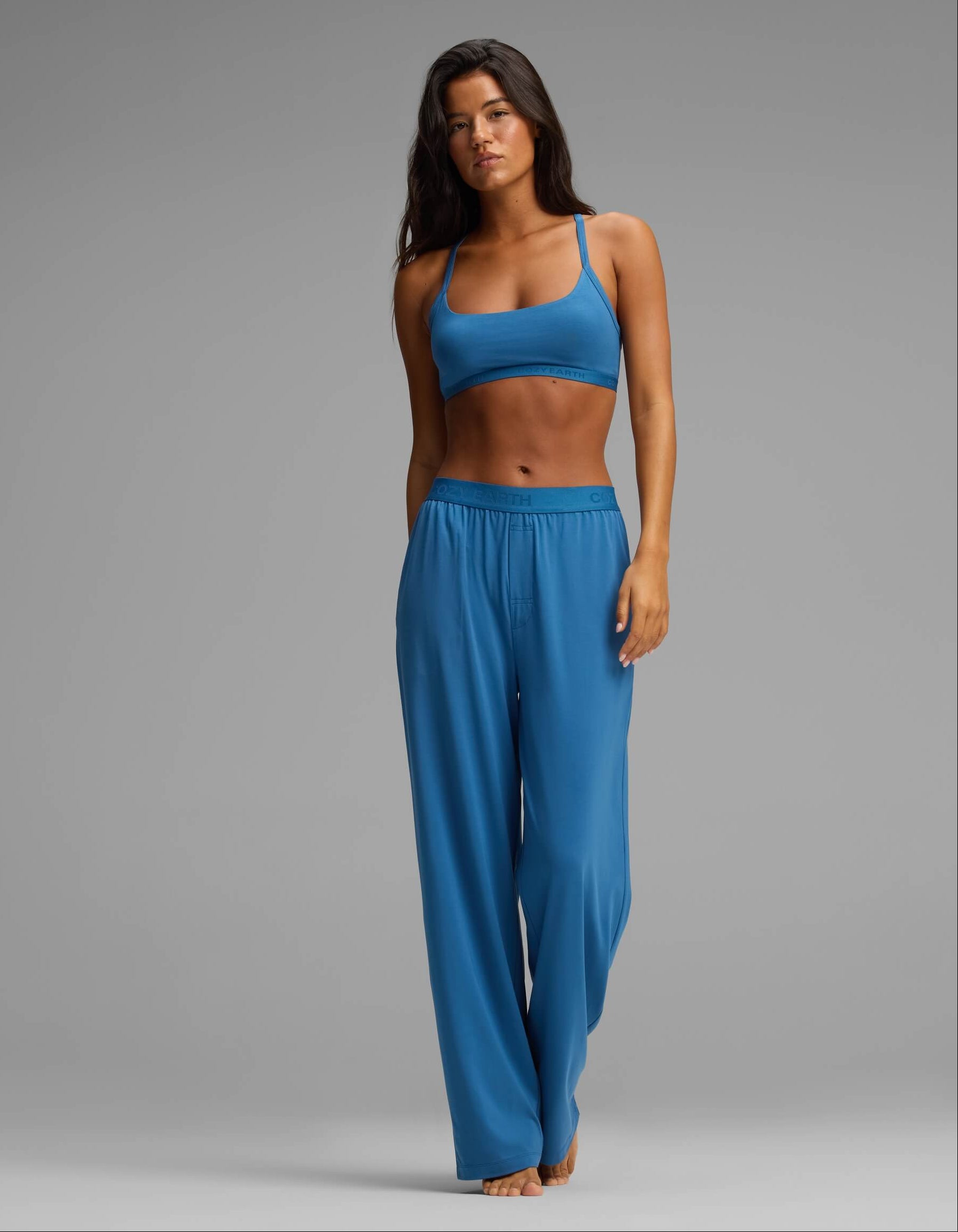 A woman stands barefoot in a studio, modeling the Cozy Earth Women’s First Layer Pajama Soft Bra & Pant Set in blue. She has long dark hair and a neutral expression, set against a plain gray background.|Color:Anchor