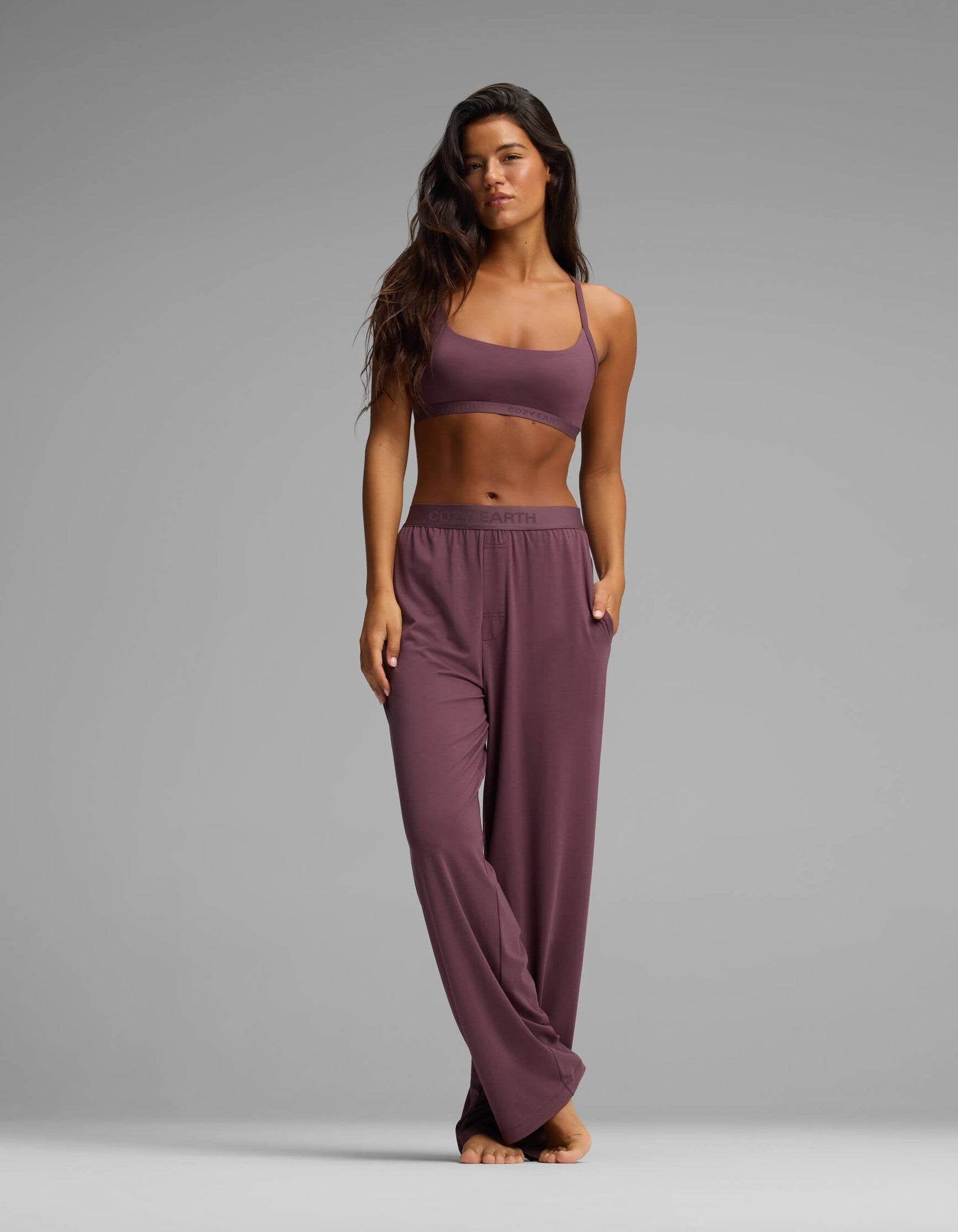 A woman stands barefoot on a gray background, wearing the Cozy Earth Women’s First Layer Pajama Soft Bra & Pant Set in mauve, with one hand in her pocket and the other by her side.|Color:Fig