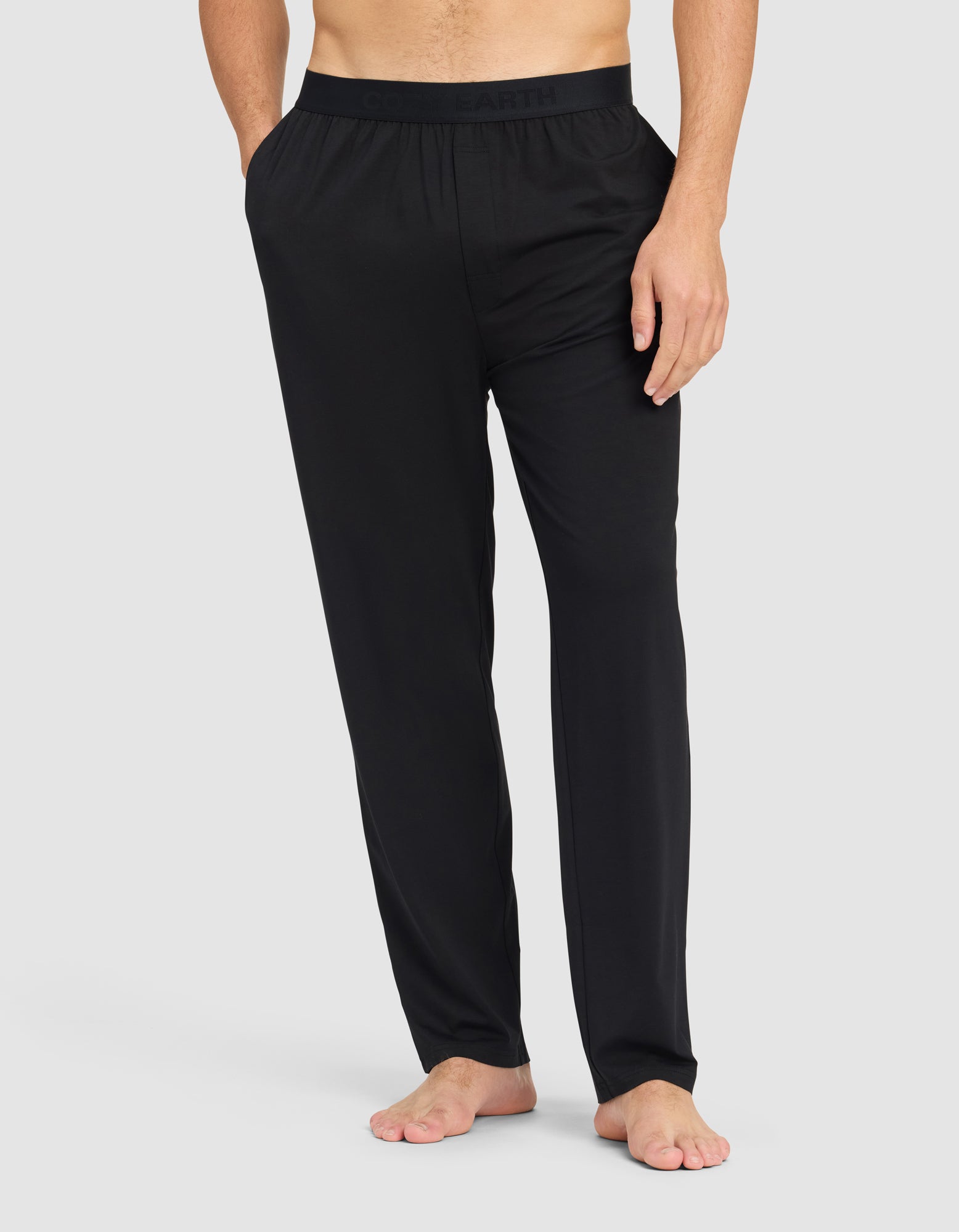 A man, with his upper body and face out of frame, stands barefoot against a light gray background wearing Cozy Earth Men's First Layer Pajama Pant—loose-fitting black pants with an elastic waistband.|Color:Jet Black
