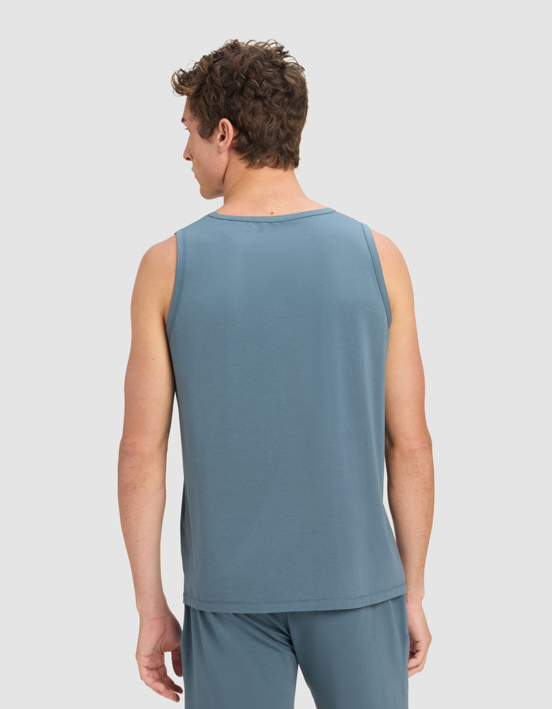 A man faces away from the camera wearing Cozy Earth’s Men's First Layer Pajama Tank in blue, paired with matching shorts. The background is plain and light-colored.