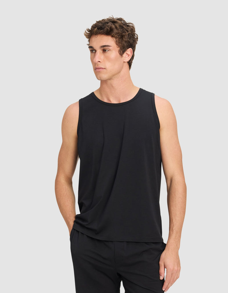 A man wears the Men's First Layer Pajama Tank by Cozy Earth and black shorts, standing with one hand in his pocket against a plain light gray background.