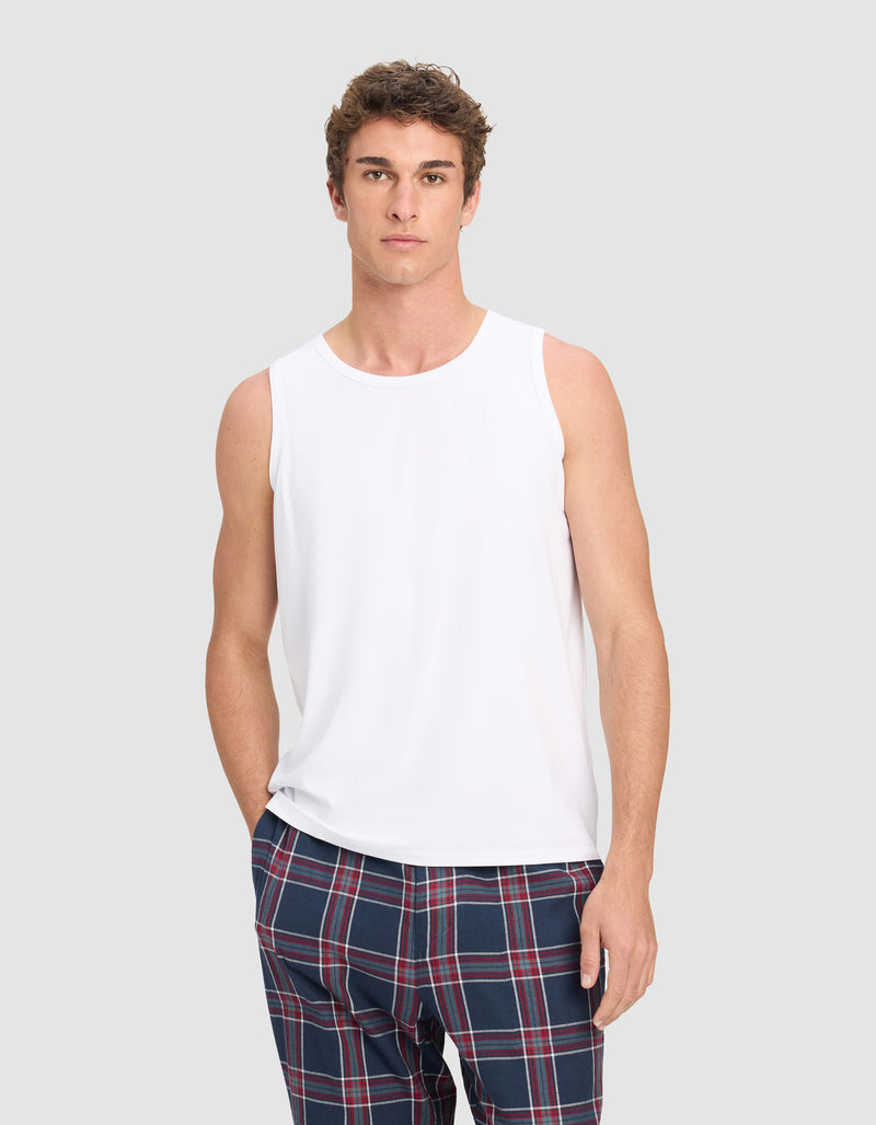 A man wears the Cozy Earth Men's First Layer Pajama Tank in white, paired with blue plaid pants, standing against a light gray background and looking directly at the camera with a neutral expression.
