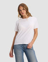 A woman with light brown hair wears the Cozy Earth Women's Go-To Tee in white and blue jeans, standing against a light gray background with one hand in her pocket and looking at the camera.|Color:White