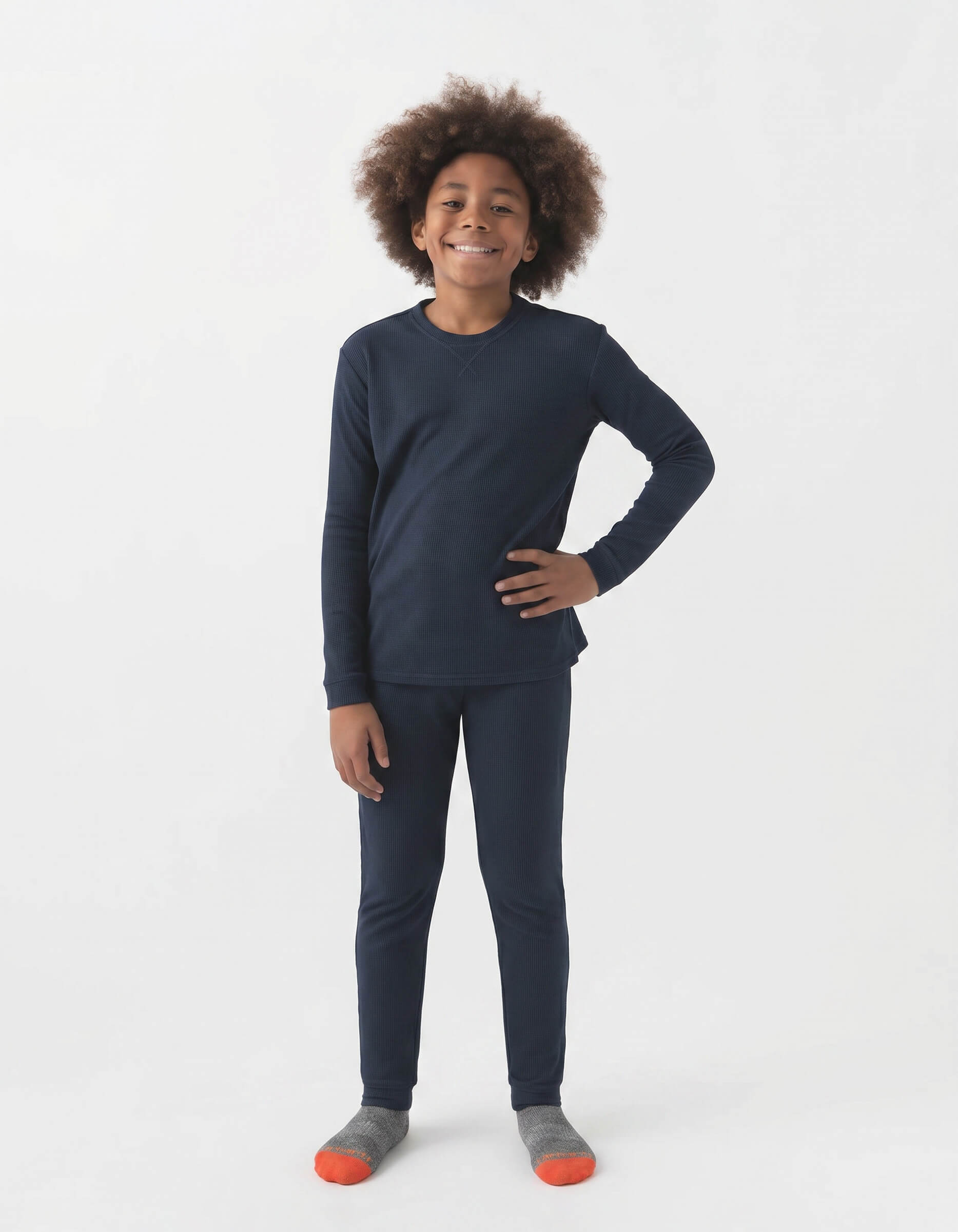 A smiling child with curly hair stands against a white background, wearing Cozy Earth's Children's Bamboo Waffle Knit Base Layer Set—navy top and pants—paired with gray and orange socks. One hand rests on their hip.