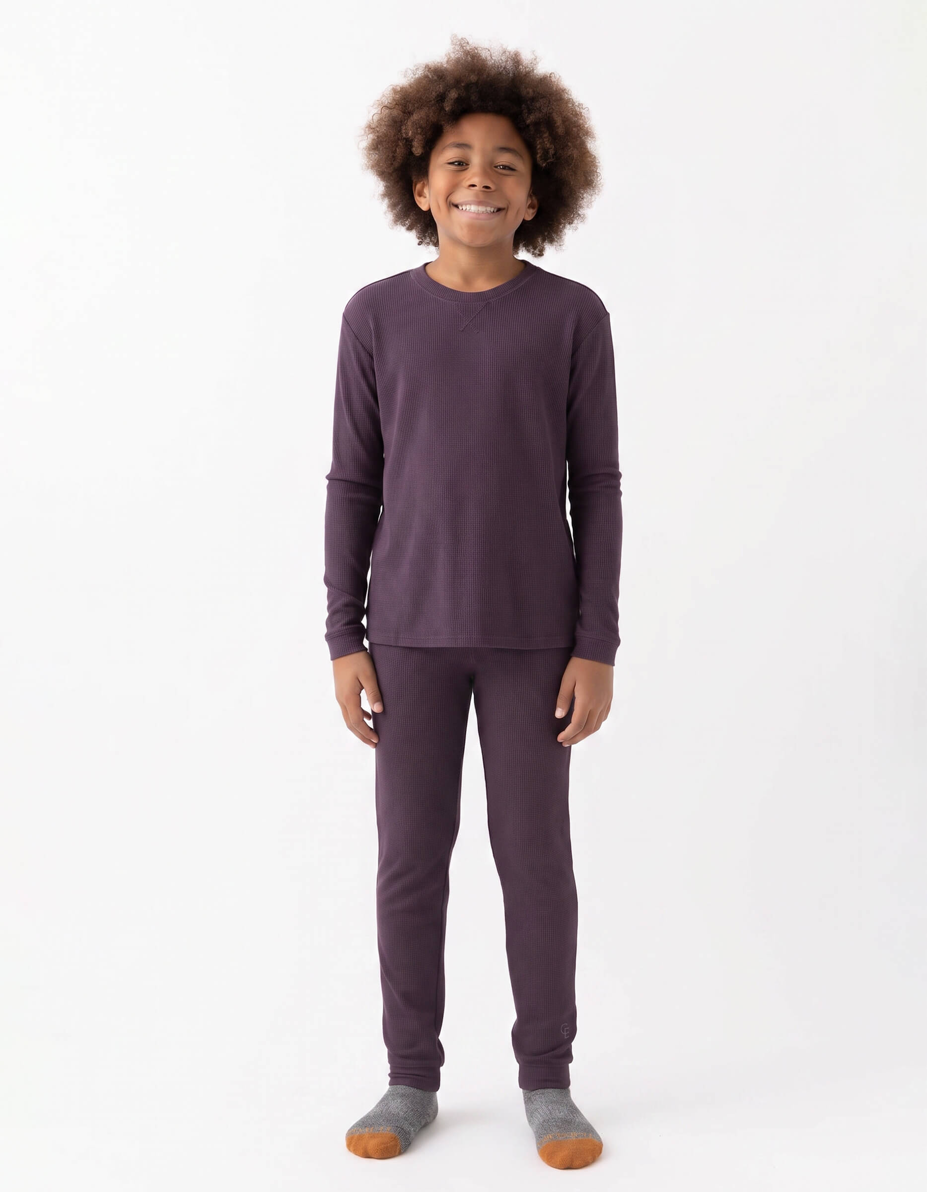 A smiling child with curly hair stands against a white background, wearing the Cozy Earth Children's Bamboo Waffle Knit Base Layer Set - Last Chance, paired with gray and orange socks.