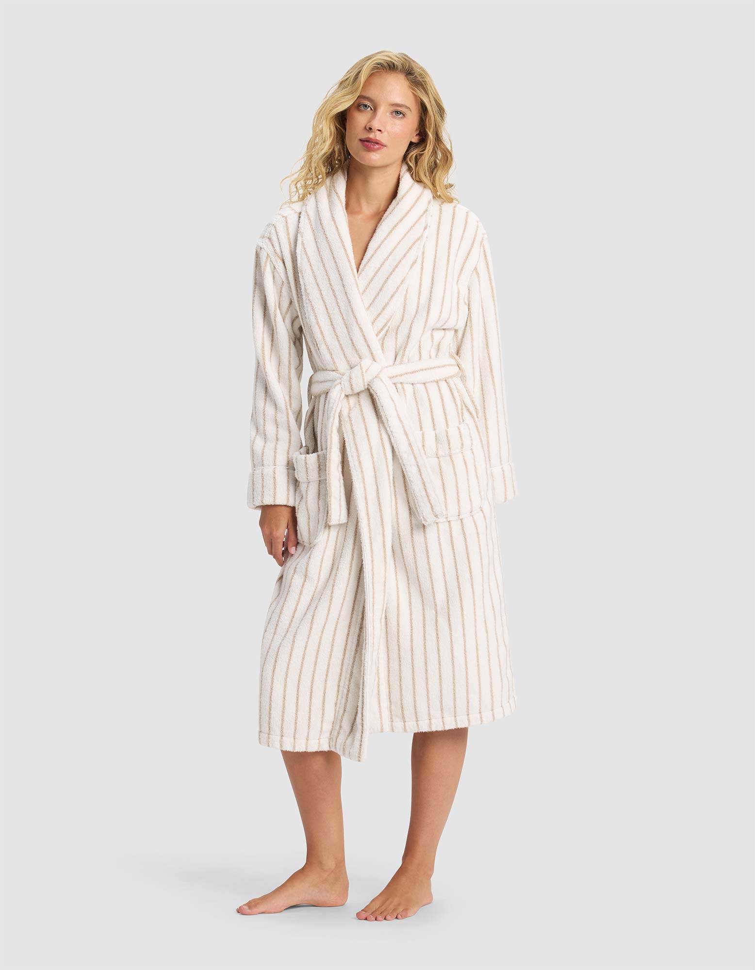 A woman stands barefoot against a light gray background, wearing the Cozy Earth Striped Luxe Bath Robe—white with beige vertical stripes and a belted waist.