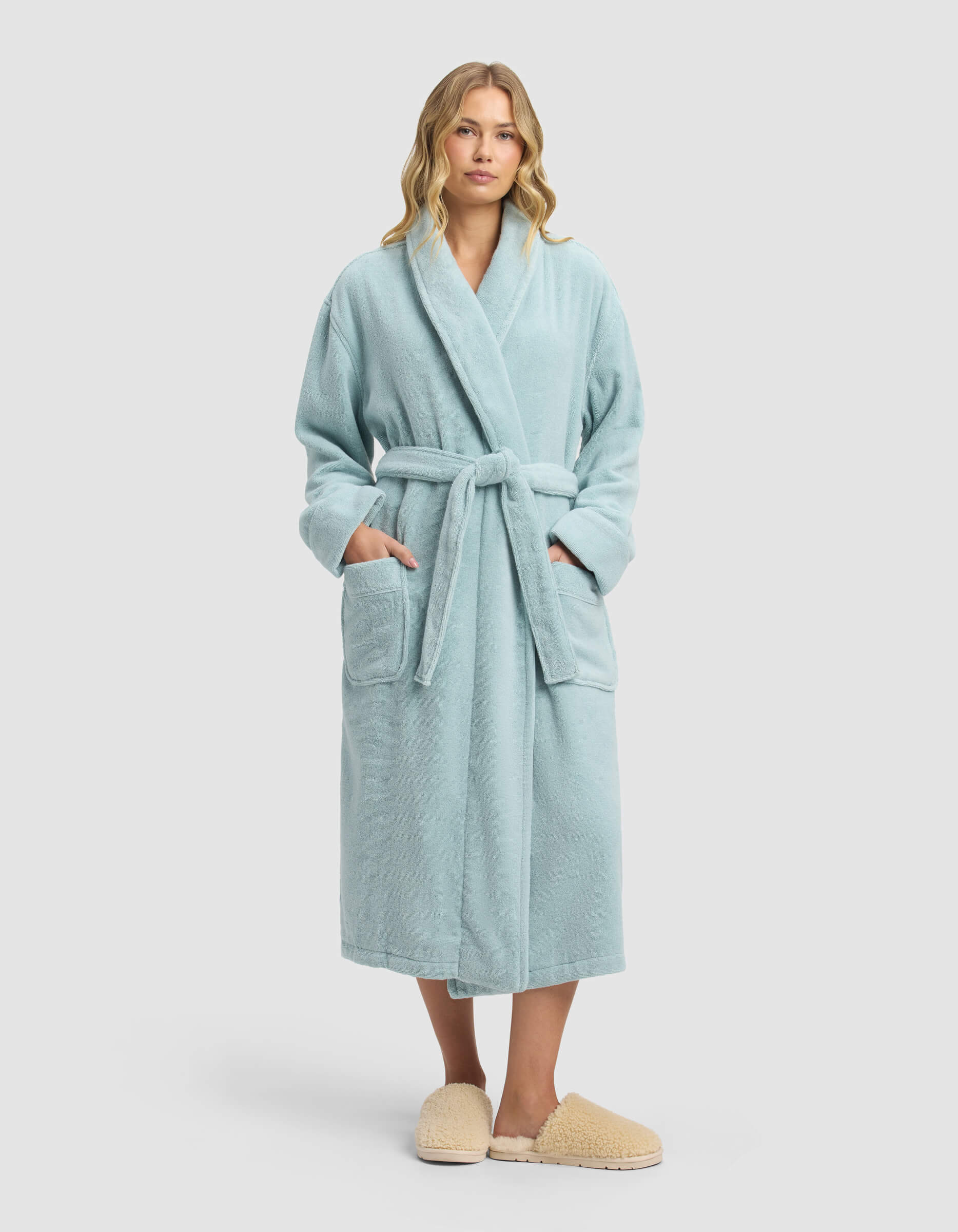 A woman stands facing forward wearing the Cozy Earth Luxe Bath Robe—light blue with a belt and large pockets—and beige slippers, set against a plain light background.|Color:Tide