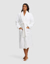 A woman with long dark hair poses barefoot against a plain light gray background, wearing the Cozy Earth Luxe Bath Robe in white. The knee-length robe features a tie belt and two front pockets.|Color:White