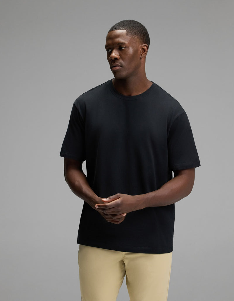 A man wearing the Cozy Earth Men’s All Day Tee in Jet Black and beige pants stands against a gray background, looking to his left with his hands loosely clasped.