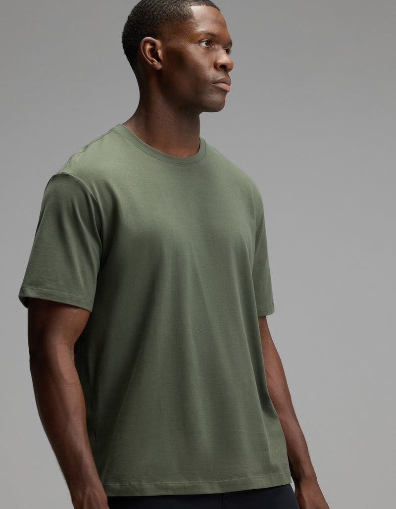 A man wearing the Cozy Earth Men’s All Day Tee in olive green stands against a neutral gray background, looking off to the side with a serious expression.
