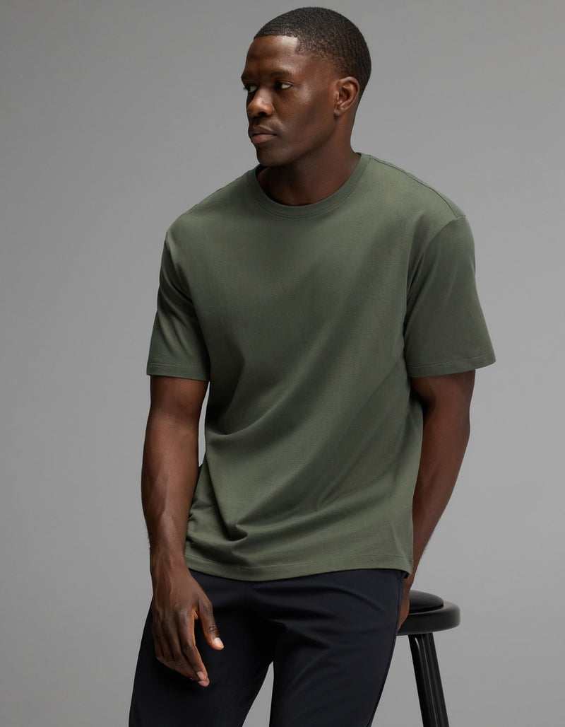 A man wearing the Cozy Earth Men’s All Day Tee in olive and black pants sits on a black stool against a plain gray background, looking to his left with a neutral expression.
