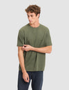 A young man with curly hair wears the Cozy Earth Men's All Day Tee in olive green and dark jeans, standing against a plain light gray background while looking down and gently pulling his sleeve. |Color:Olive Heather