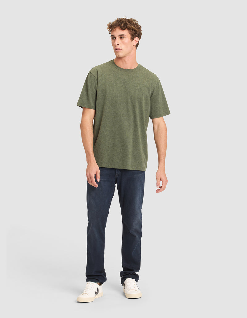 A man with short curly hair and a neutral expression wears the Cozy Earth Men's All Day Tee in olive green, paired with dark blue jeans and white sneakers, standing against a plain light gray background with his left hand relaxed by his side.