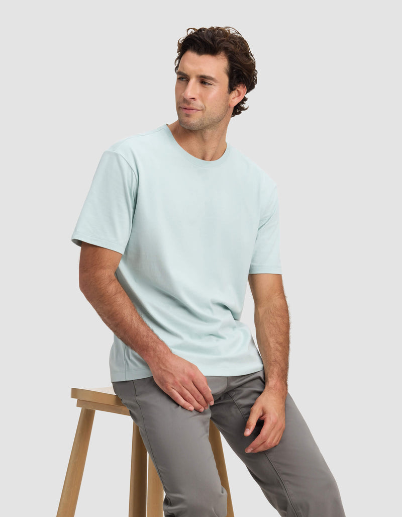 A man with short brown hair wears the Cozy Earth Men's All Day Tee in light blue and gray pants, seated on a wooden stool against a plain light gray background, looking slightly left with a relaxed expression.