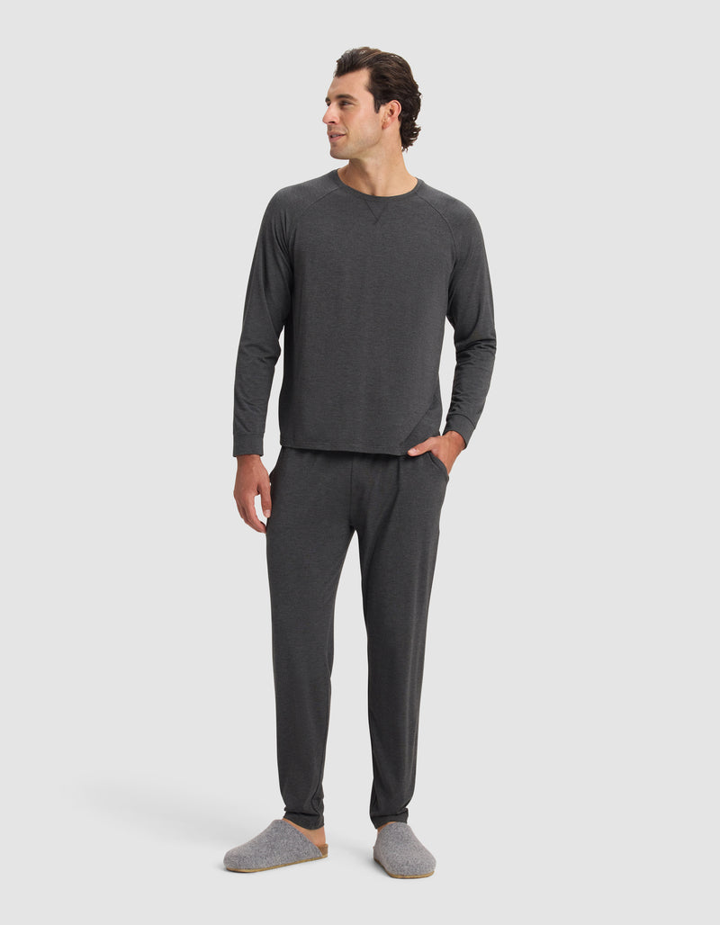 A man with short dark hair smiles in a plain white setting, wearing Cozy Earth Men's Bamboo Stretch-Knit Pajama Pants and matching dark gray long-sleeve top, paired with gray slippers and one hand in his pocket.