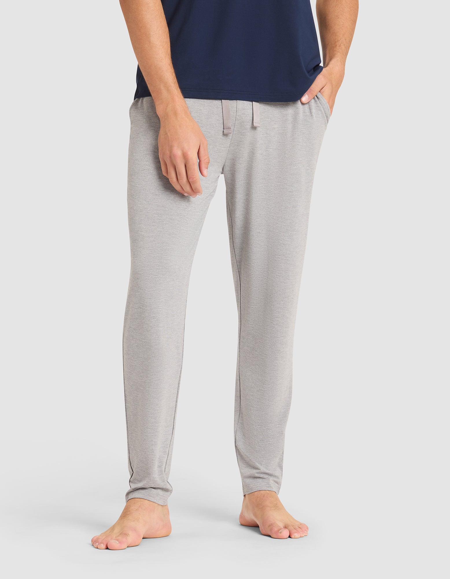 A person is barefoot, wearing Cozy Earth Men's Bamboo Stretch-Knit Pajama Pant in light gray with hands in the pockets, paired with a navy blue shirt. The background is plain white. |Color:Heather Grey