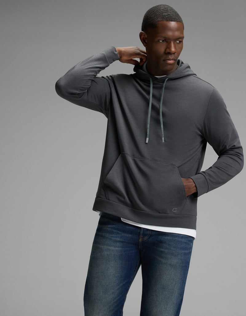A man models the Cozy Earth Men’s Brushed Bamboo Hoodie in dark gray with blue jeans, standing against a plain gray background and gazing to the side while resting one hand on the back of his neck.