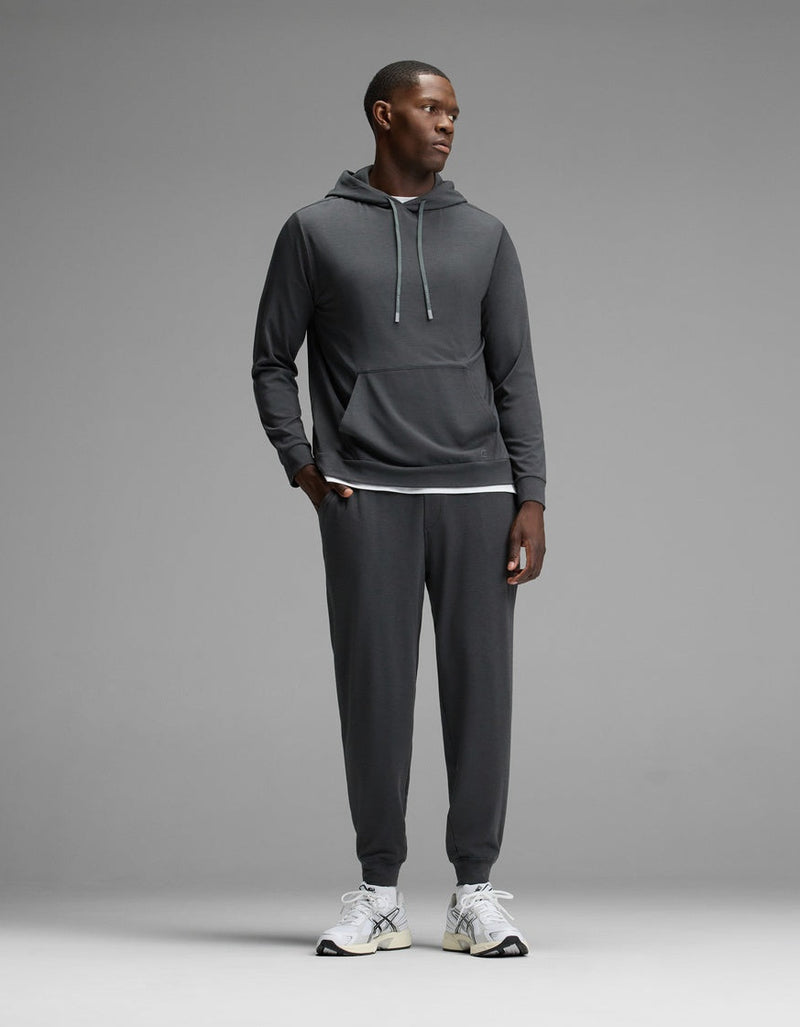 A man stands against a grey background, wearing the Cozy Earth Men’s Brushed Bamboo Hoodie & Jogger Set in Charcoal, paired with white athletic shoes. He looks to the side with his hands by his sides.