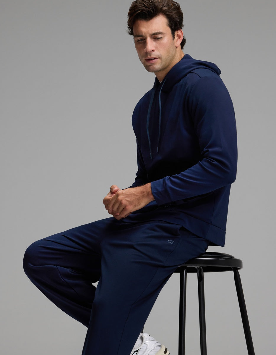 A man wears the Cozy Earth Men’s Brushed Bamboo Hoodie & Jogger Set in navy blue, sitting on a black stool against a plain gray background, looking slightly downward with his hands resting near his lap.