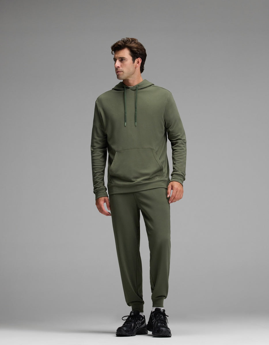 A man models the Cozy Earth Men’s Brushed Bamboo Hoodie & Jogger Set in olive, standing against a plain gray background. He looks slightly to his left with his hands relaxed at his sides, completing the look with black sneakers.