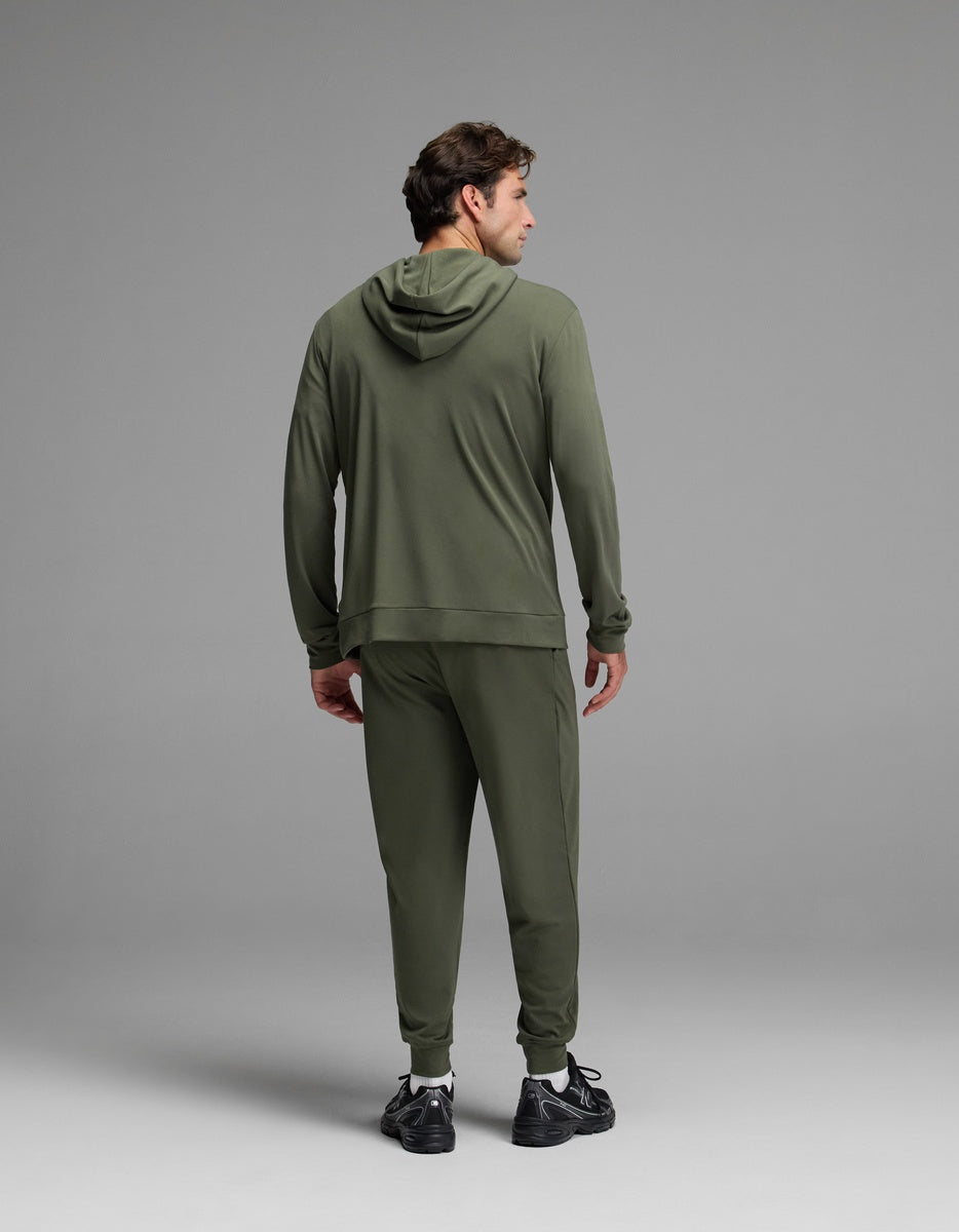 A man stands facing away, wearing Cozy Earth’s Men’s Brushed Bamboo Hoodie & Jogger Set in olive green with black sneakers, set against a plain gray background.