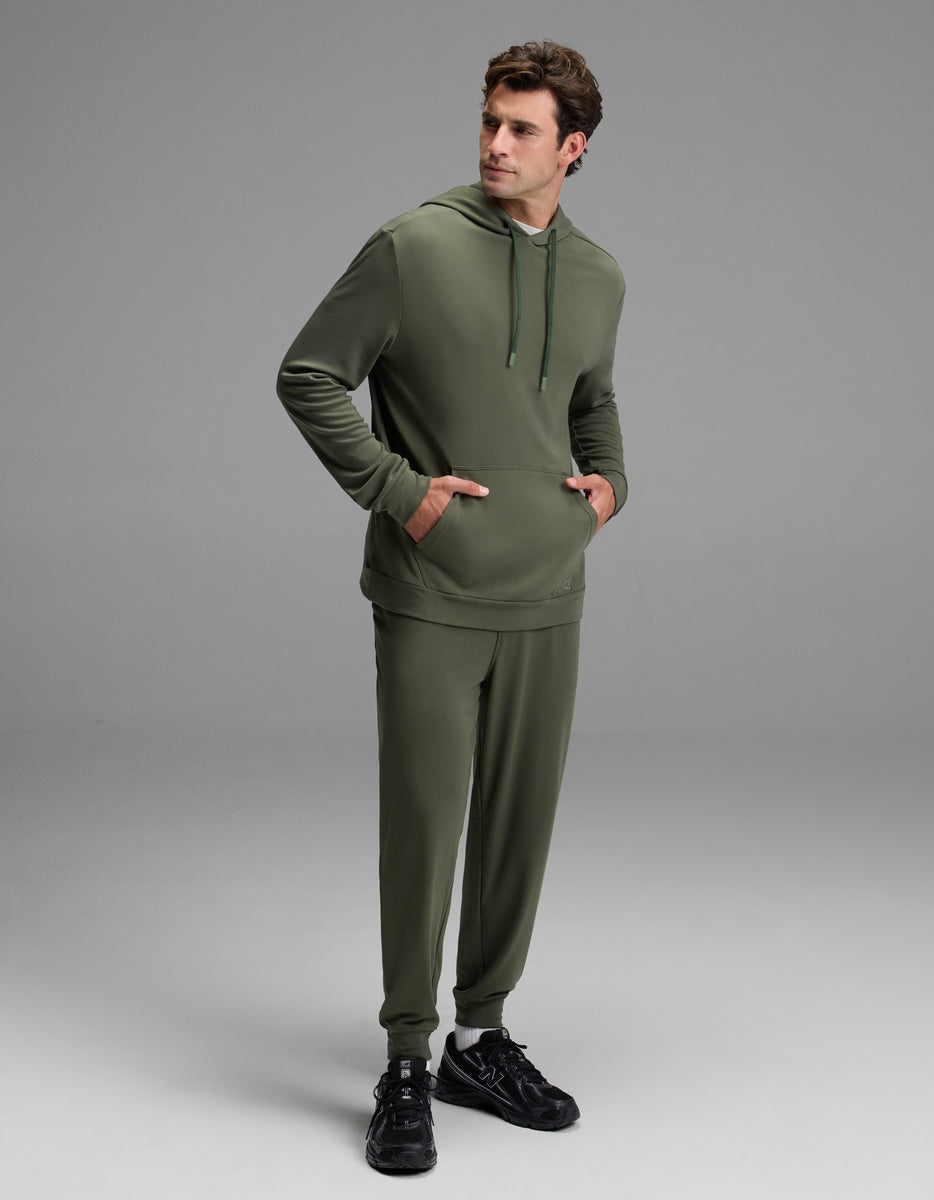 A man dressed in the Cozy Earth Men’s Brushed Bamboo Hoodie & Jogger Set in olive green stands with his hands in his pockets, looking to the side. He completes the look with black sneakers against a plain light gray background.