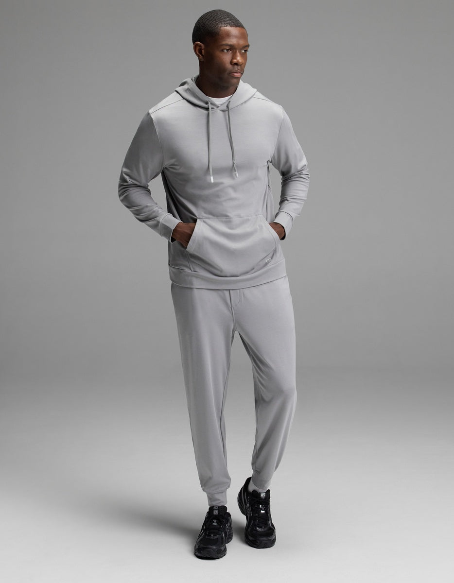 A man wearing the Cozy Earth Men’s Brushed Bamboo Hoodie & Jogger Set in Stone stands against a plain gray background, looking to his left with his hands in the hoodie pocket and black sneakers on.|Color:Stone